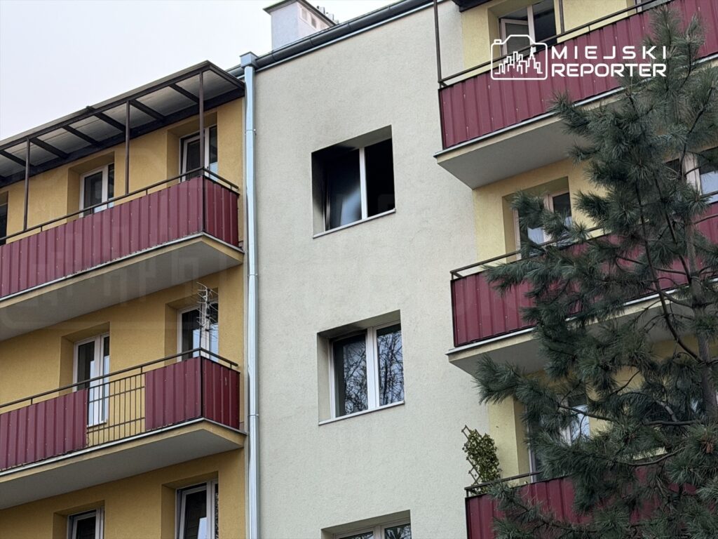 Budynek mieszkalny z żółtą elewacją i czerwonymi balkonami, z widocznymi oknami i drzewem w tle.