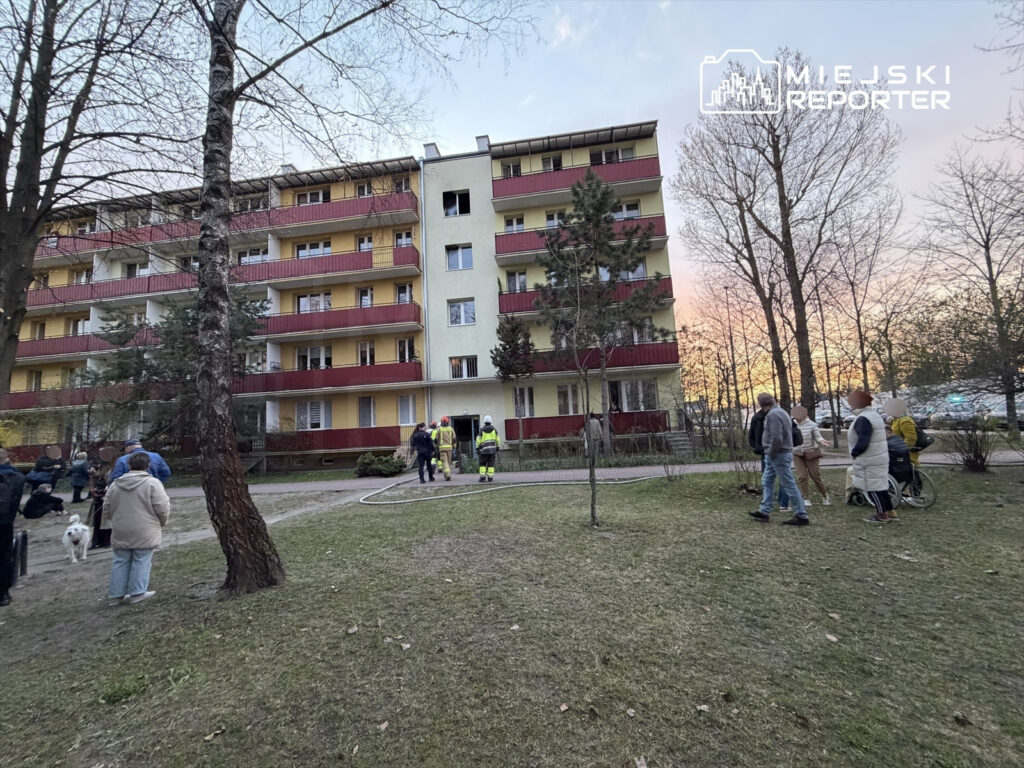 Grupa ludzi obserwuje działania strażaków przy bloku mieszkalnym w czasie zachodu słońca.