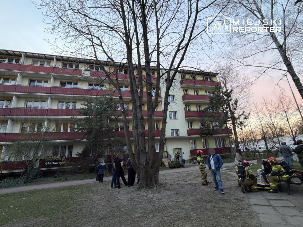 Grupa strażaków udziela pomocy w pobliżu bloku mieszkalnego, obok stoją zaniepokojeni mieszkańcy.