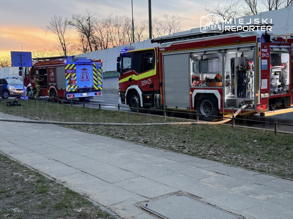 W tle zachód słońca, strażacy pracują przy dwóch wozach strażackich, jeden z otwartą komorą z wyposażeniem.