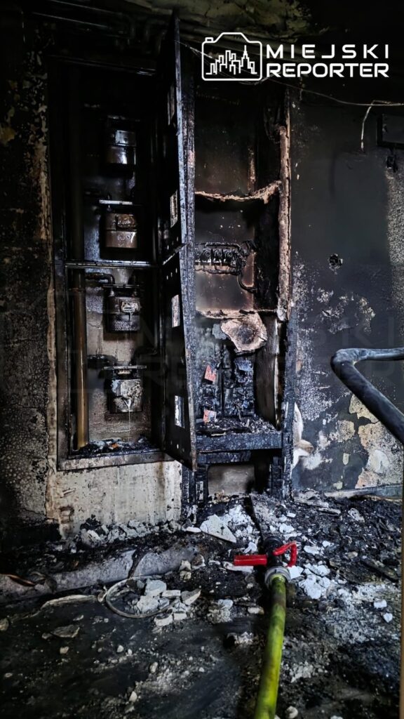 Zniszczona ściana z otwartą szafą elektryczną, pokrytą sadzą, oraz wężykiem gaśniczym leżącym na zniszczonym podłożu.