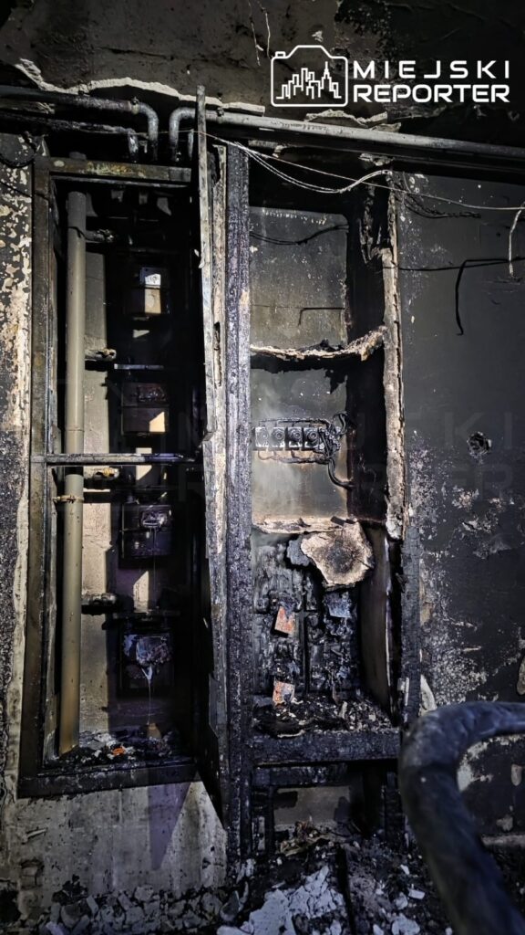 Zniszczona instalacja elektryczna z popiołem i sadzą w ciemnym pomieszczeniu, z widocznymi uszkodzeniami i zniszczeniami.