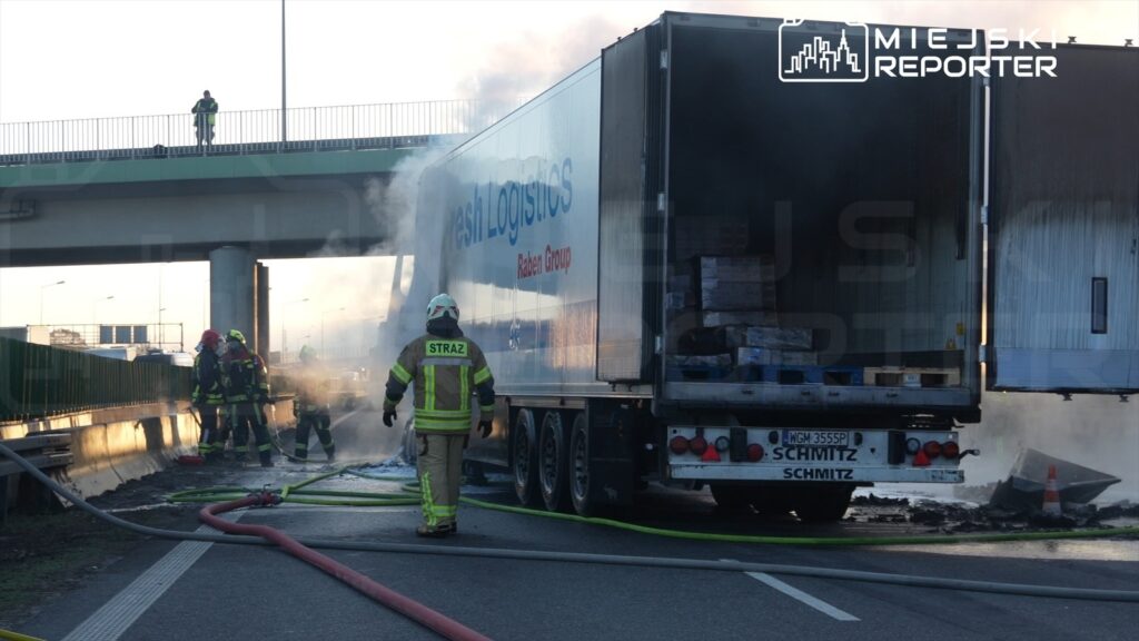 Strażacy gaszą ogień w uszkodzonej ciężarówce na autostradzie, obok rozrzucone węże gaśnicze i dym.