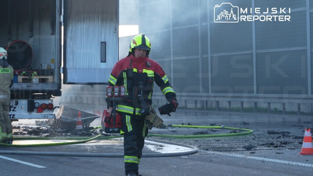 Strażak w specjalistycznym stroju niesie sprzęt gaśniczy obok uszkodzonego pojazdu ciężarowego na drodze.