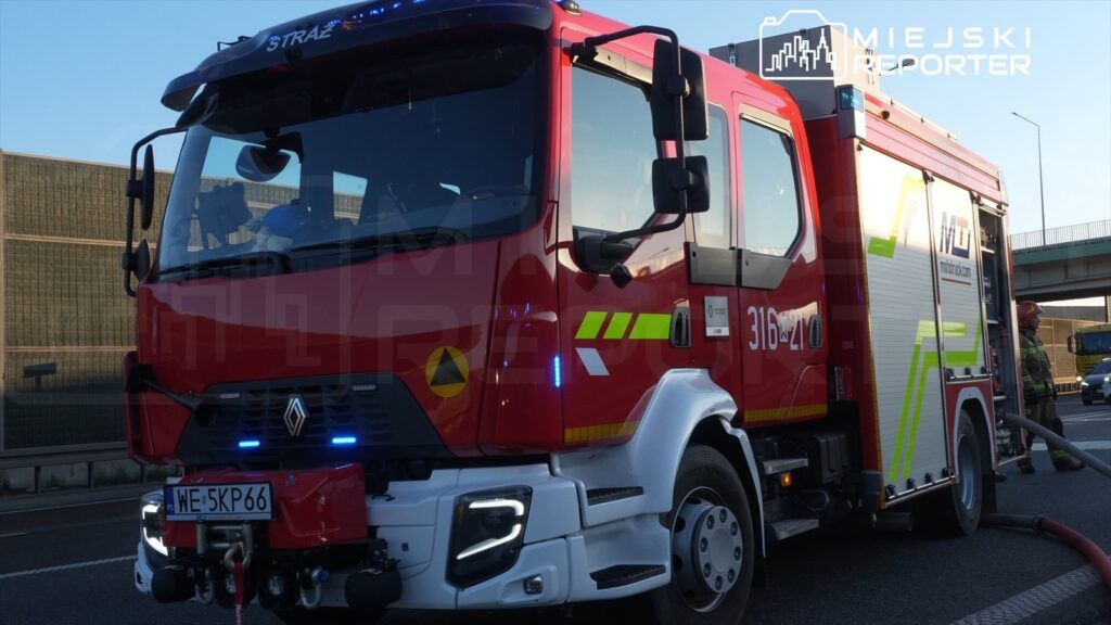 Czerwony wóz strażacki z niebieskimi światłami parkuje przy drodze, obok stoi strażak z wężem gaśniczym.