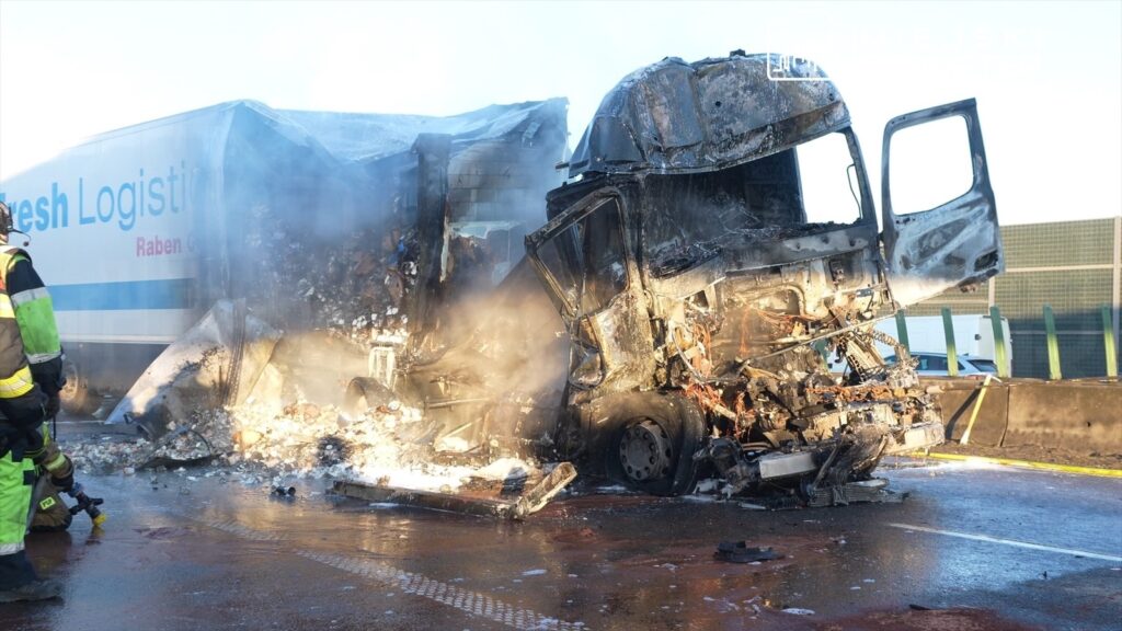 Strażak w zielonym mundurze gasi pożar zniszczonej ciężarówki na drodze, w tle widoczne są pojazdy i dym.