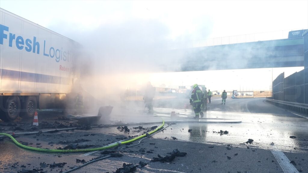 Strażacy gaszą pożar przy ciężarówce na drodze, w tle widoczny most i dym unoszący się w powietrzu.
