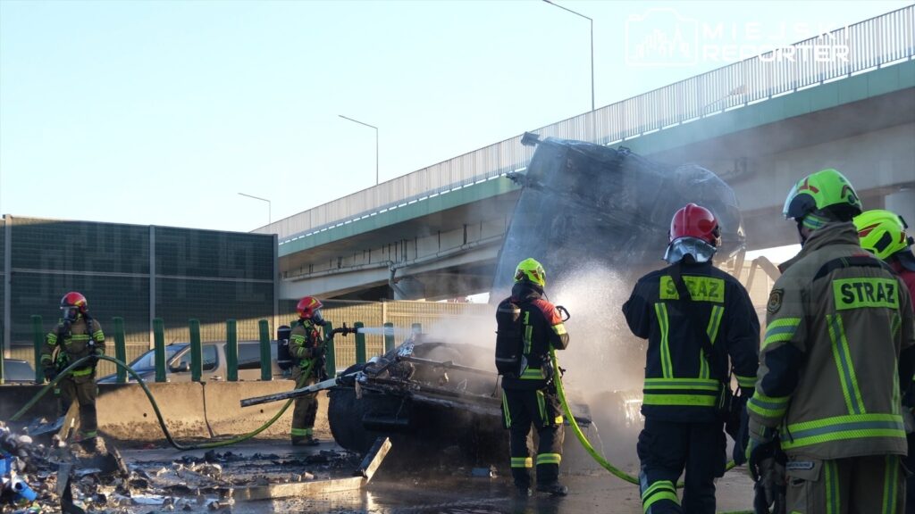 Strażacy gaszą płonący wrak pojazdu na drodze pod mostem, wokół widoczne zniszczone elementy.