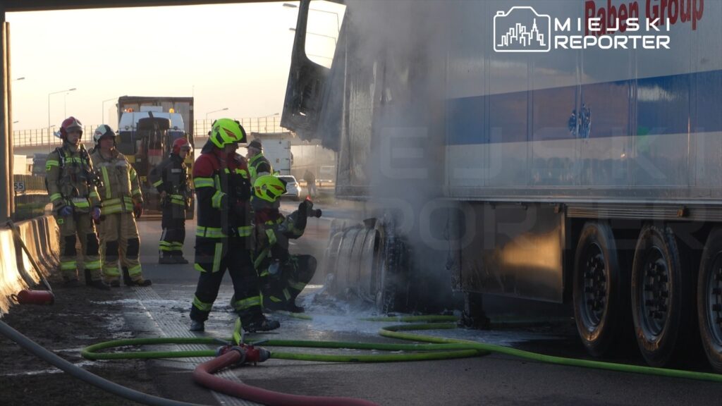 Grupa strażaków gasi pożar w naczepie ciężarówki na drodze, otoczeni sprzętem ratunkowym i dymem.