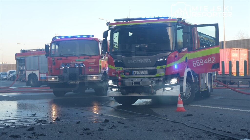 Dwa wozy strażackie z włączonymi sygnałami świetlnymi na drodze, w tle widoczne kontenery.