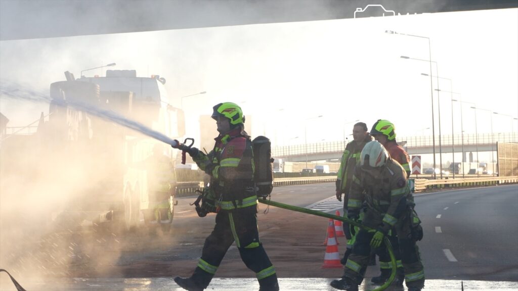 Strażacy w hełmach gaszą pożar przy drodze, otoczeni gęstym dymem, z włączonymi sygnałami i sprzętem.
