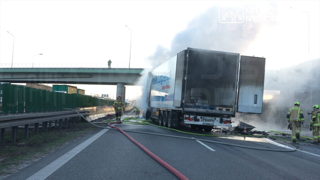 Strażacy gaszą pożar ciężarówki na autostradzie, w tle widoczny dym i uszkodzona naczepa.