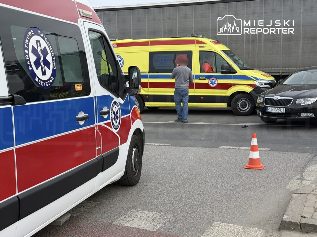 Ambulans z czerwonymi i niebieskimi oznaczeniami stoi obok żółtego karetki, a mężczyzna w szarej koszuli rozmawia z ratownikami.