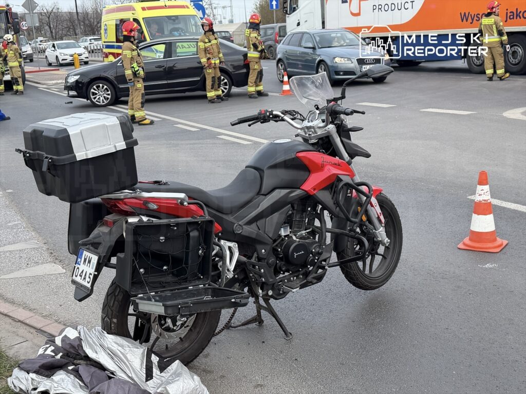 Motocykl z bagażnikiem stoi na jezdni obok miejsca wypadku, gdzie strażacy pracują nad zabezpieczeniem terenu.