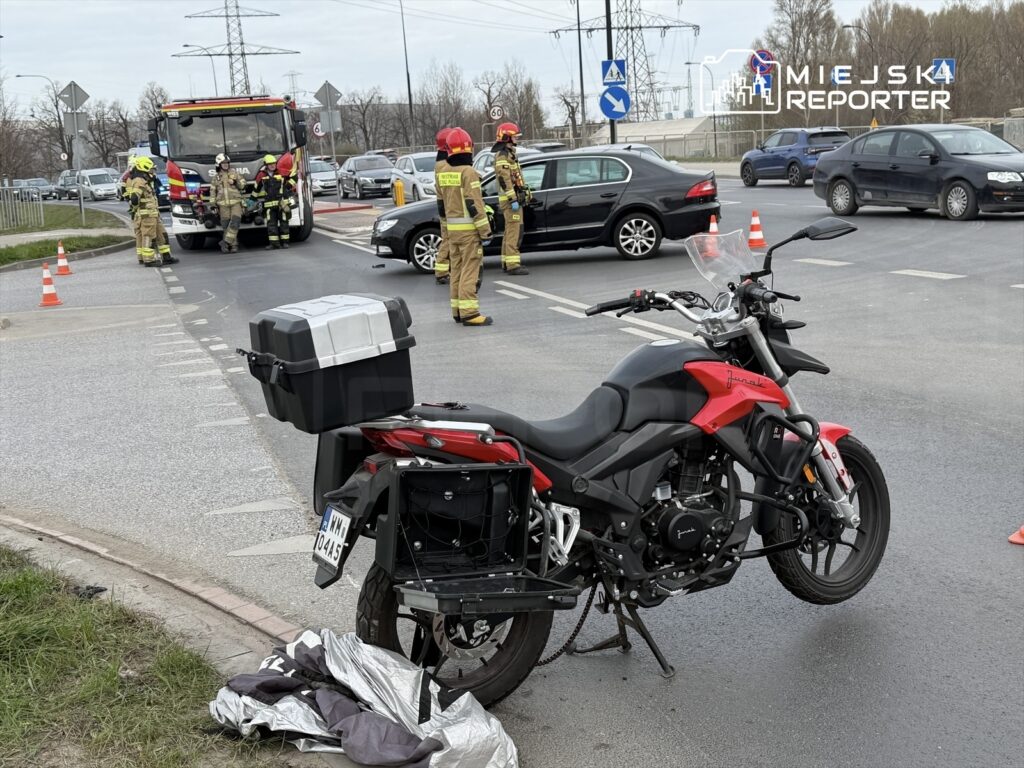 Motocykl leży na poboczu drogi, obok którego pracują strażacy i policjanci, a w tle widać ruch samochodowy.