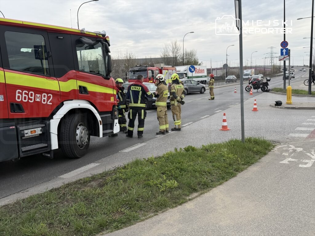 Grupa strażaków w mundurach omawia sytuację obok wozu strażackiego na ruchliwej drodze.