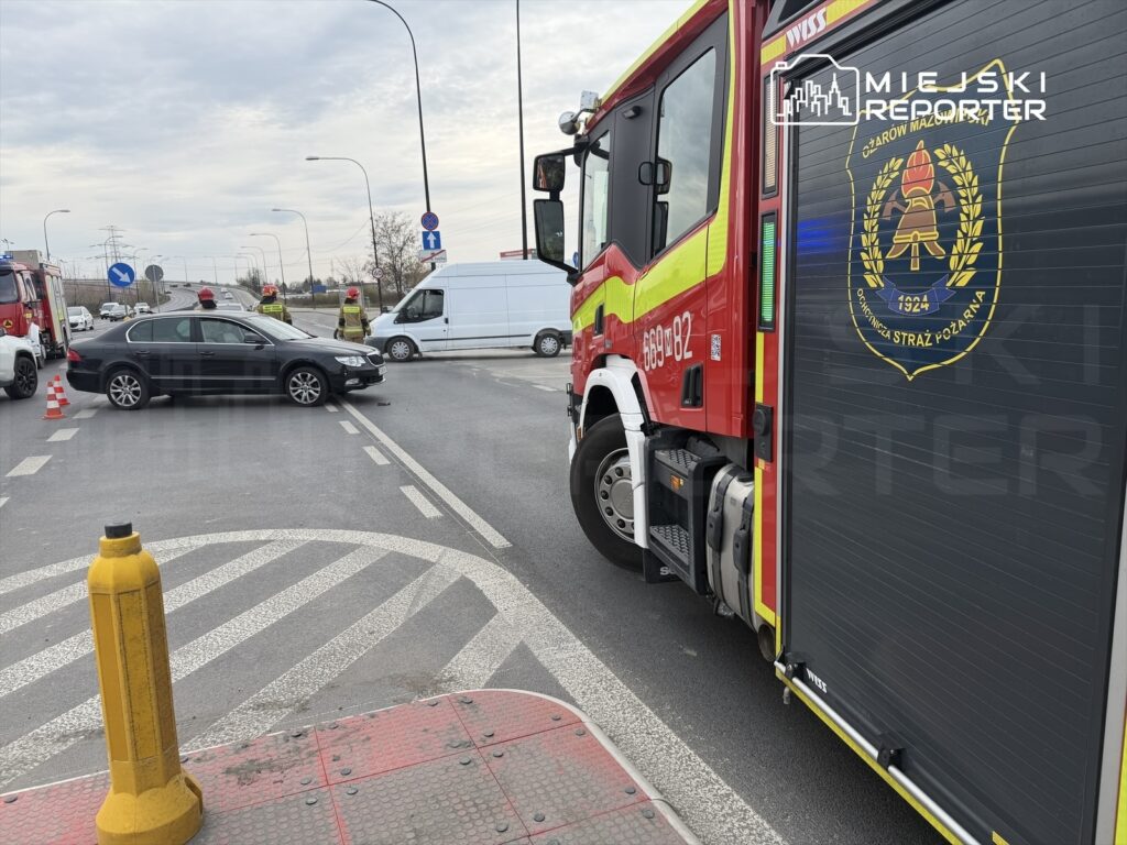 Czarny samochód osobowy stoi na skrzyżowaniu, obok strażacy w kaskach zabezpieczających, a w tle widać wóz strażacki.