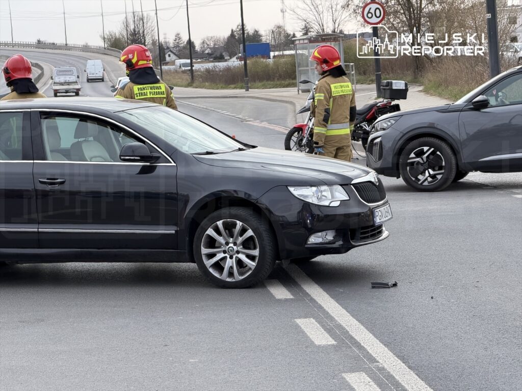 Czterech strażaków w hełmach interweniuje przy zderzeniu dwóch samochodów na drodze, obok znaku ograniczenia prędkości.