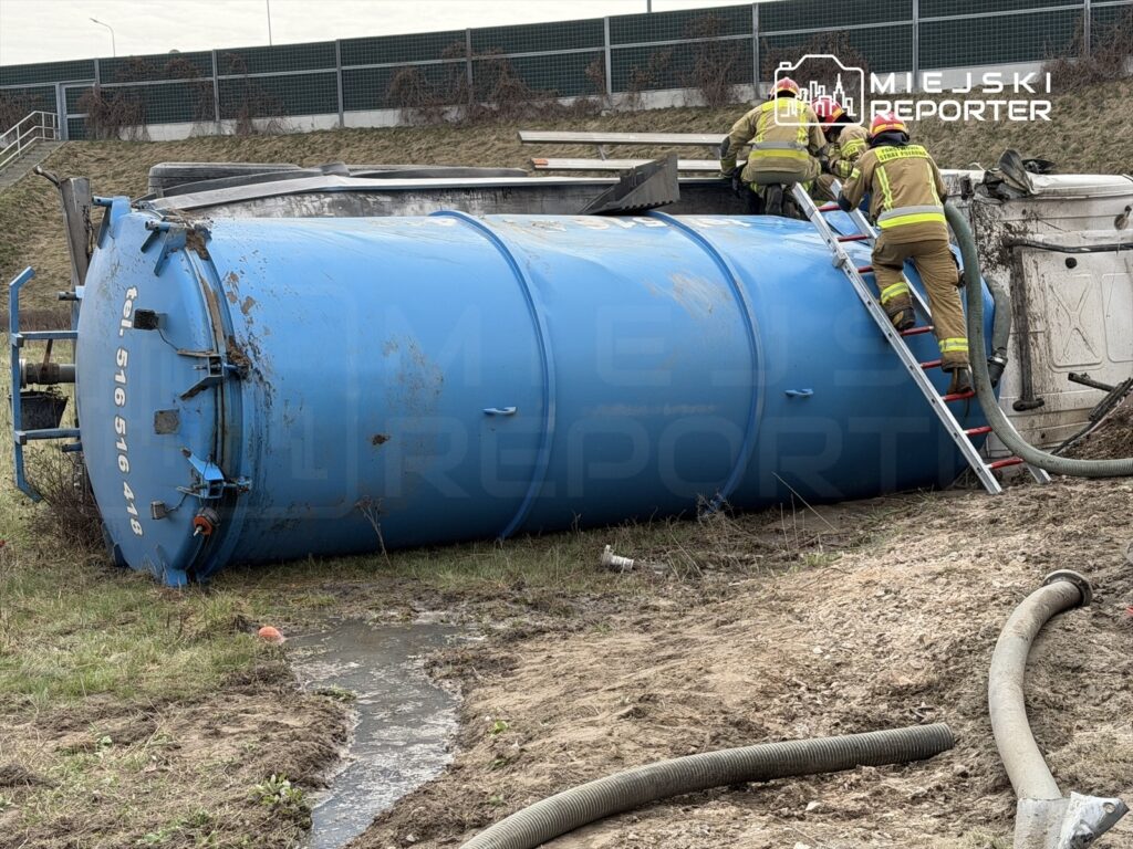 Dwaj strażacy podchodzą do przewróconego niebieskiego zbiornika, używając drabiny w terenie zielonym.