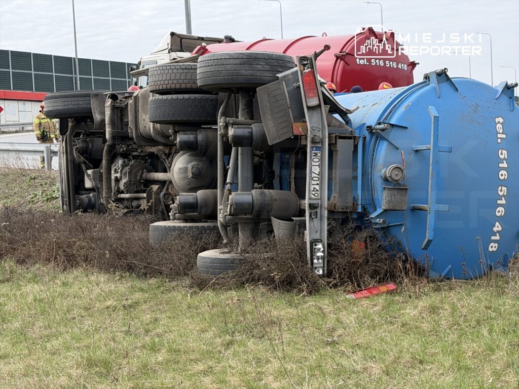 Czerwony ciężarowy pojazd leży na boku obok drogi, a obok niego stoi strażak w mundurze.