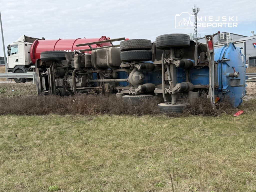 Niebieski samochód ciężarowy leży na boku na poboczu drogi, otoczony trawą i krzewami.