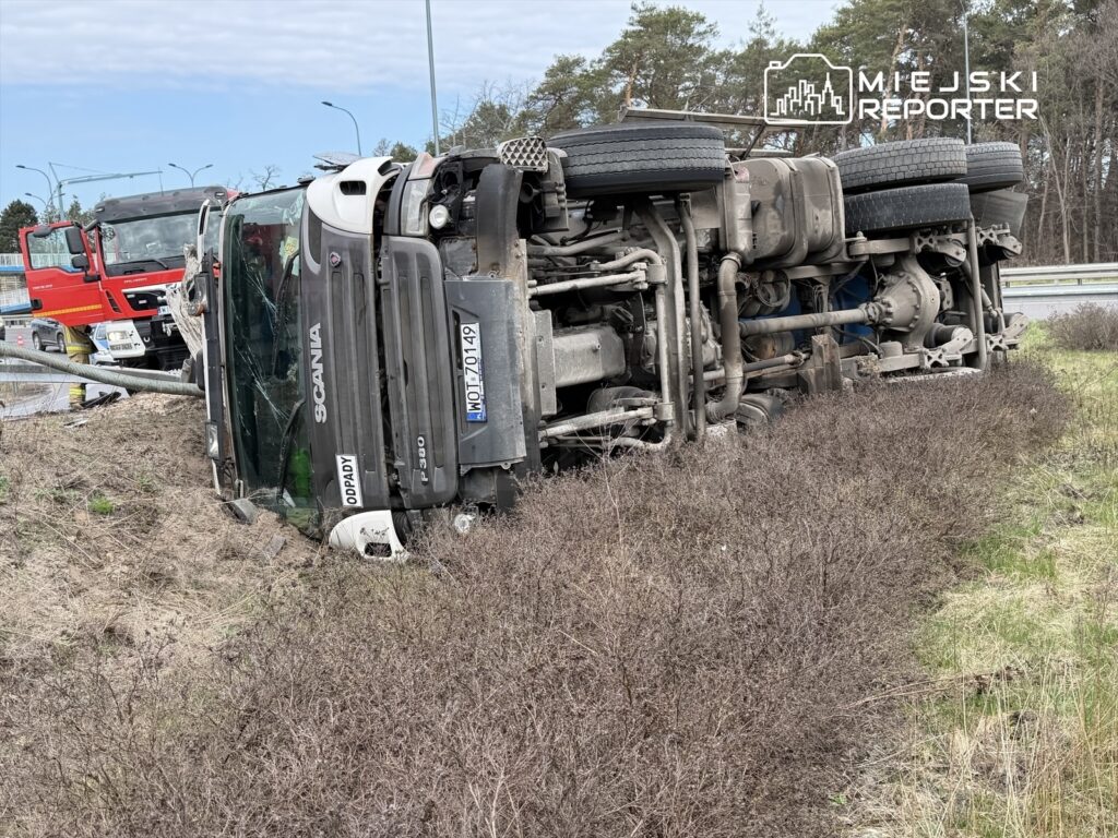 Czarny ciężarówka przewrócona na bok w rowie przy drodze, w tle widoczny wóz strażacki.