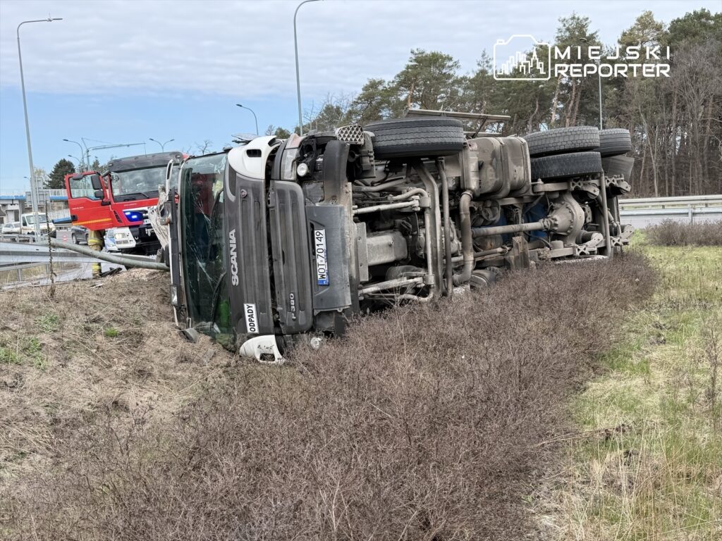 Czarna ciężarówka marki Scania leży na boku na poboczu drogi, obok widać interweniujące pojazdy służb ratunkowych.