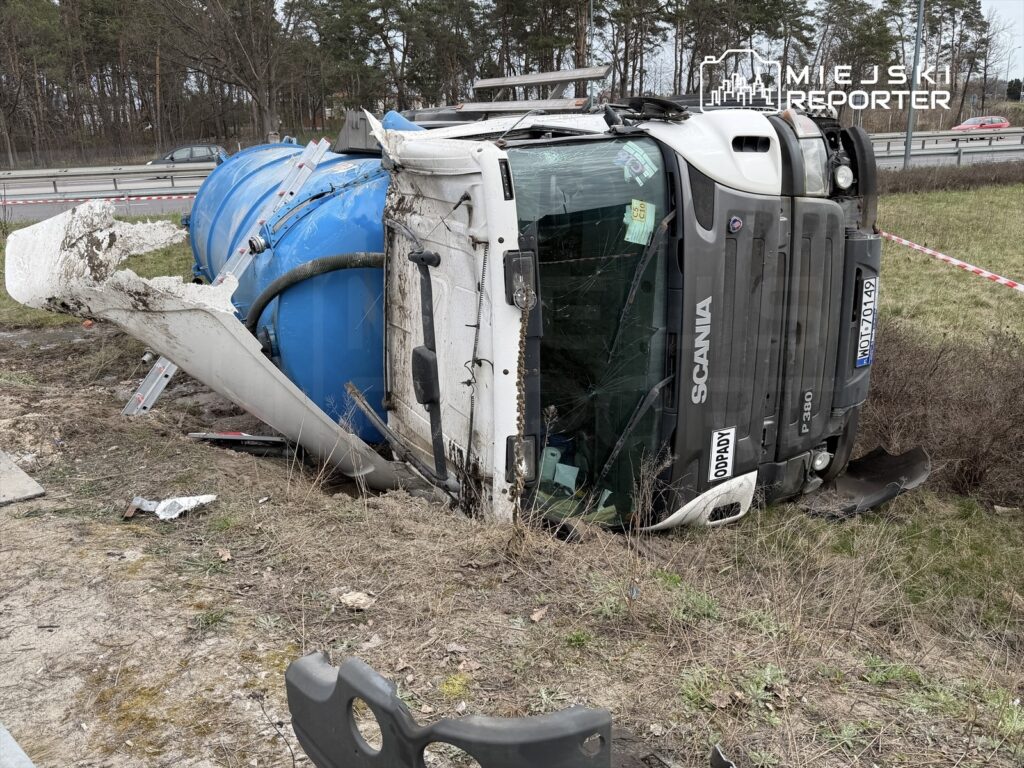 Cysterna przewrócona na bok na poboczu drogi, w otoczeniu drzew i oznakowania drogowego.