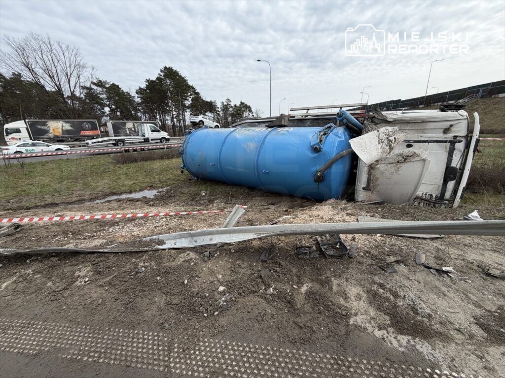 Przewrócony niebieski zbiornik na drodze, obok uszkodzone barierki, w tle przejeżdżające ciężarówki.