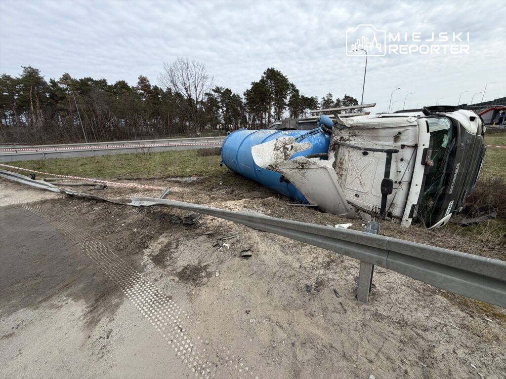 Przewrócony ciężarowy pojazd z niebieskim zbiornikiem leży na poboczu drogi, obok uszkodzonego bariery ochronnej.