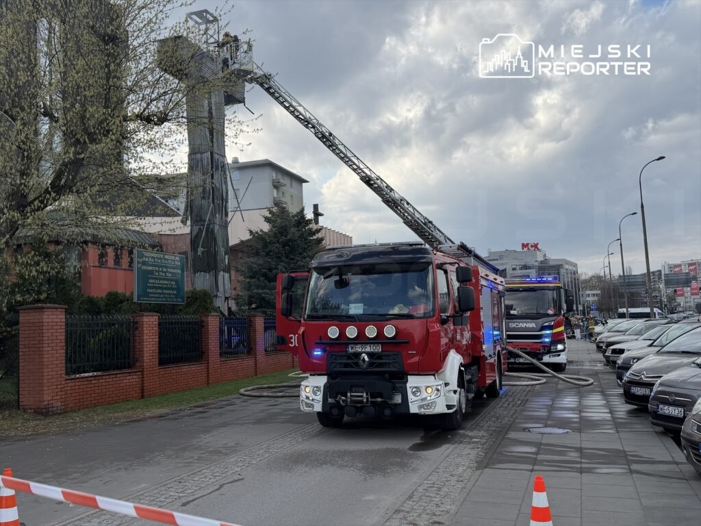 Wozidła strażackie na ulicy, jeden z pojazdów z drabiną, strażacy pracujący na wysokości przy budynku.