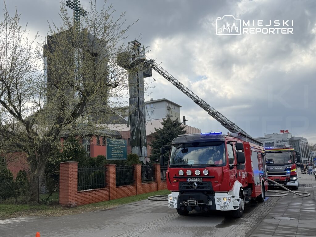 Czerwony wóz strażacki z włączonymi sygnałami świetlnymi stoi na ulicy, obok wznoszącej się wieży strażackiej.