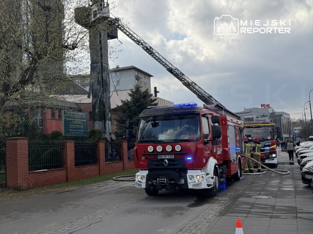Czerwony wóz strażacki z podnośnikiem i wężem strażackim zaparkowany przed budynkiem, obok strażacy w hełmach.