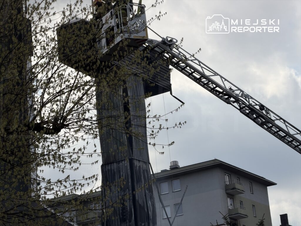 Strażacy na podnośniku wykonują prace na wysokości w pobliżu budynku mieszkalnego, w tle widoczna chmurna niebo.