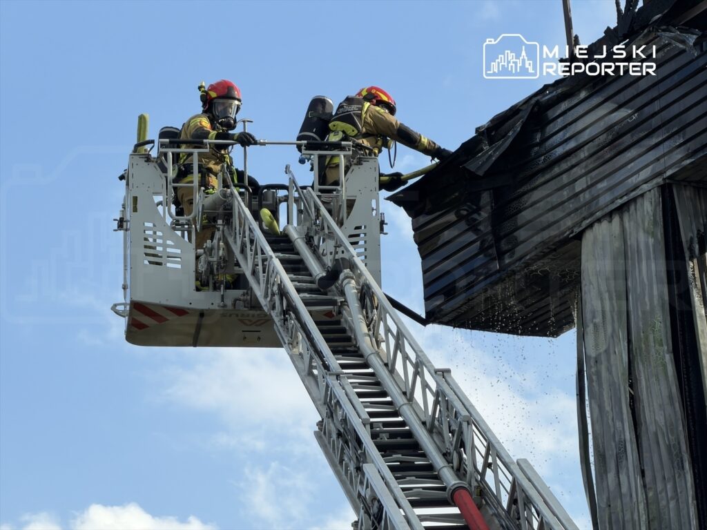 Strażacy w hełmach używają podnośnika, aby gasić pożar na dachu budynku, w tle błękitne niebo.