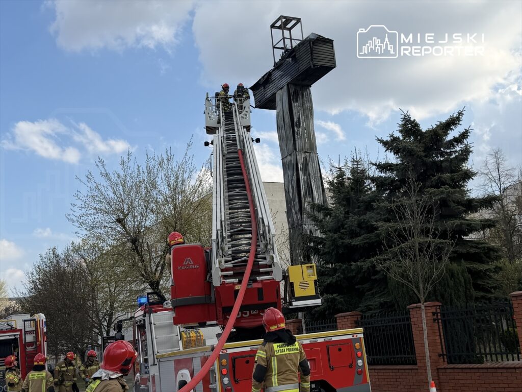 Strażacy na drabinie wysięgnikowej gaszą pożar na dachu budynku, w otoczeniu drzew i chmur.