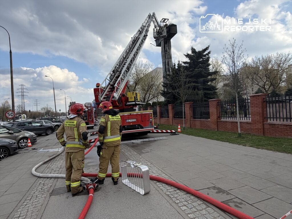 Dwóch strażaków w mundurach, z wężem strażackim, obserwuje działanie wozu strażackiego z podnośnikiem.