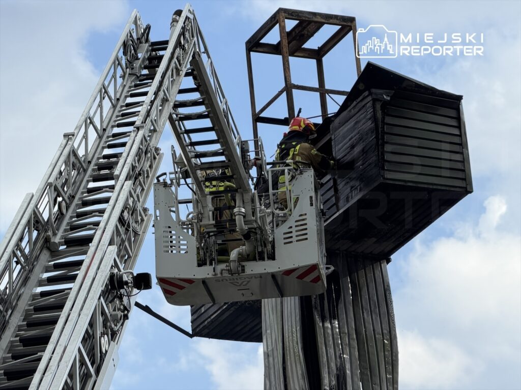Strażacy w specjalistycznym wozie strażackim przeszukują zniszczoną konstrukcję na wysokości, w tle niebo.