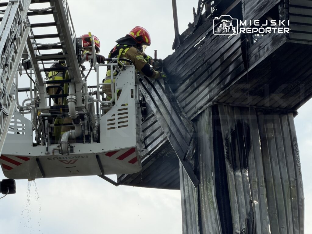 Dwaj strażacy w hełmach gaszą pożar z wysokości drabiny, polewając wodą z węża czarną, uszkodzoną konstrukcję.