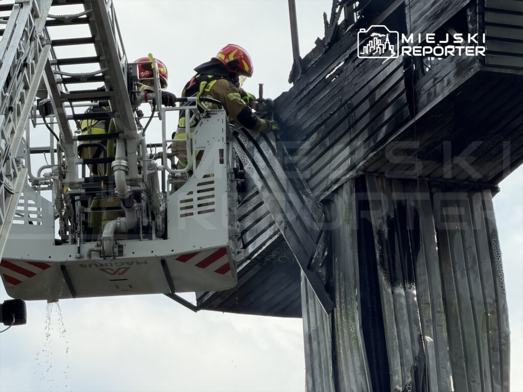 Dwaj strażacy w hełmach gaszą pożar na wysokości, korzystając z drabiny strażackiej.