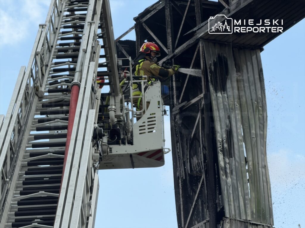 Strażak w hełmie używa węża gaśniczego do gaszenia ognia na uszkodzonym budynku.