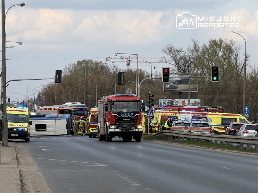 W miejscu wypadku drogowym leży przewrócony pojazd, a w pobliżu pracują strażacy i ratownicy medyczni.