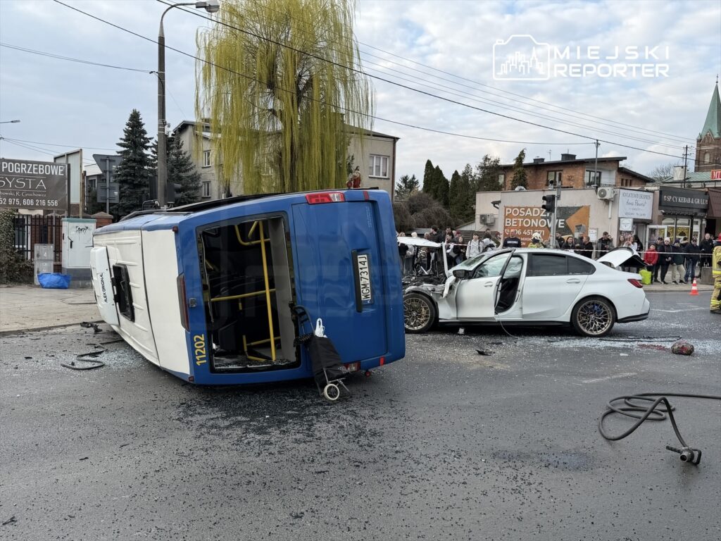 Na drodze leży przewrócony niebieski autobus, obok uszkodzony biały samochód osobowy, a wokół zbierają się gapie.