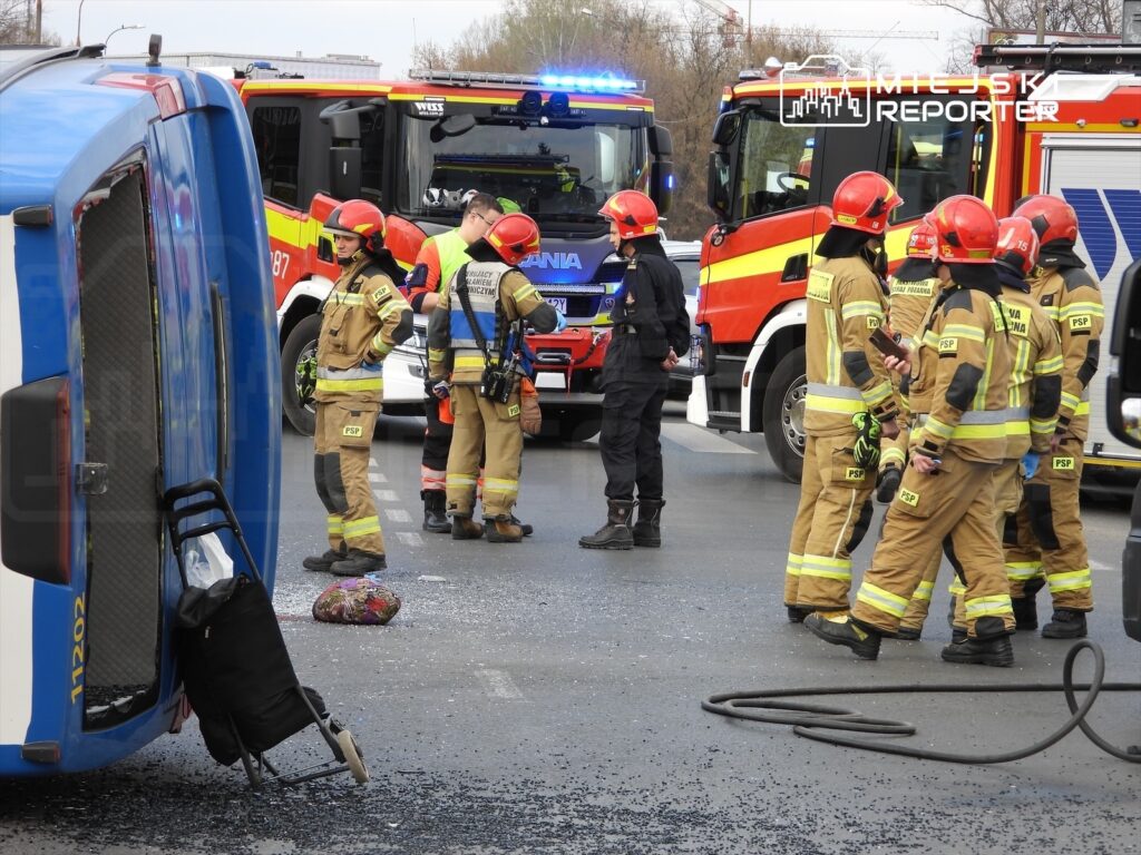 Grupa strażaków w mundurach z odblaskami pracuje na miejscu wypadku drogowego, obok przewróconego pojazdu.