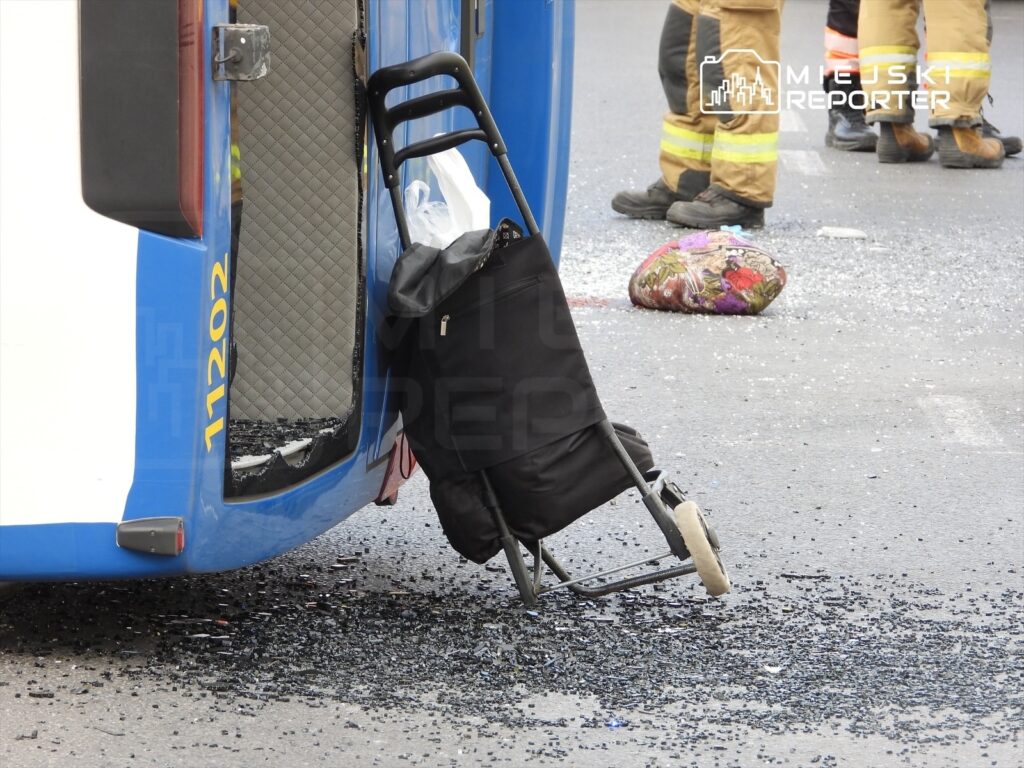 Wózek dziecięcy oparty o niebieski pojazd, obok rozbite szkło na jezdni, w tle strażacy w mundurach.