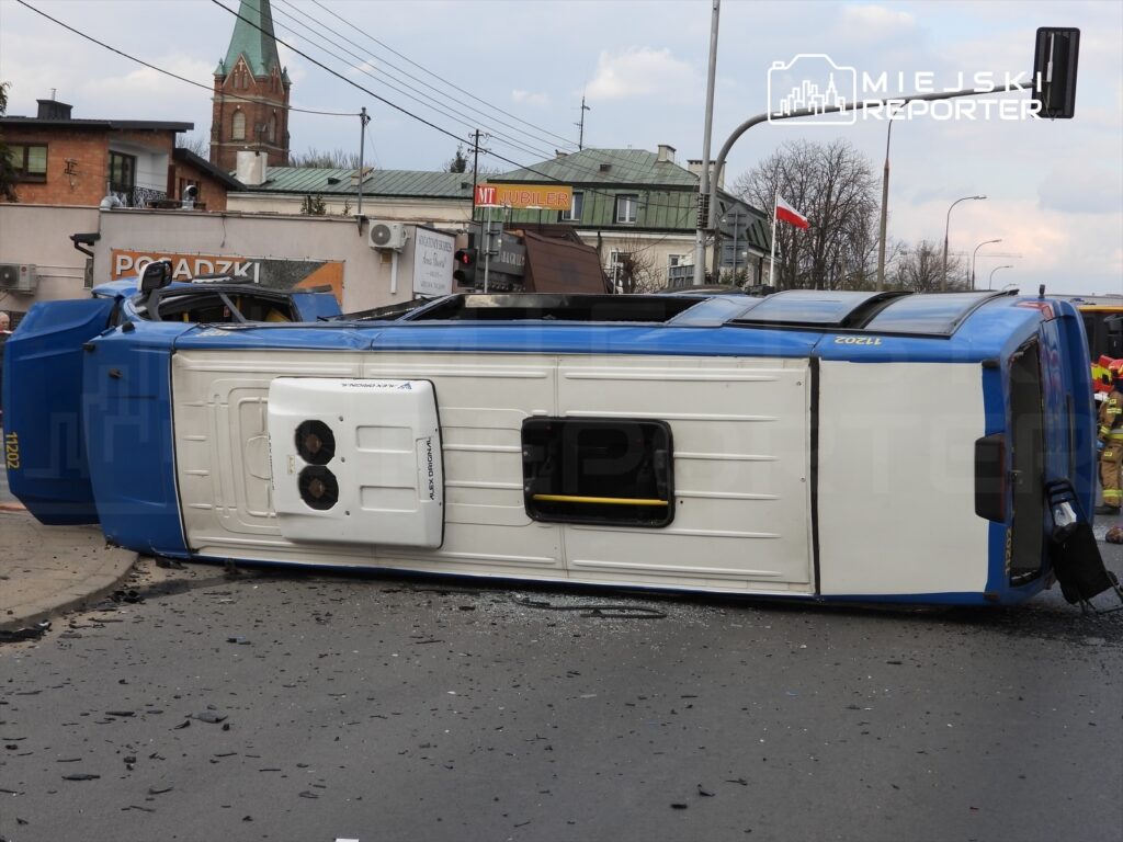 Niebieski pojazd dostawczy leży na boku na jezdni, otoczony odłamkami szkła i resztkami wypadku. W tle widoczny jest kościół oraz budynki.