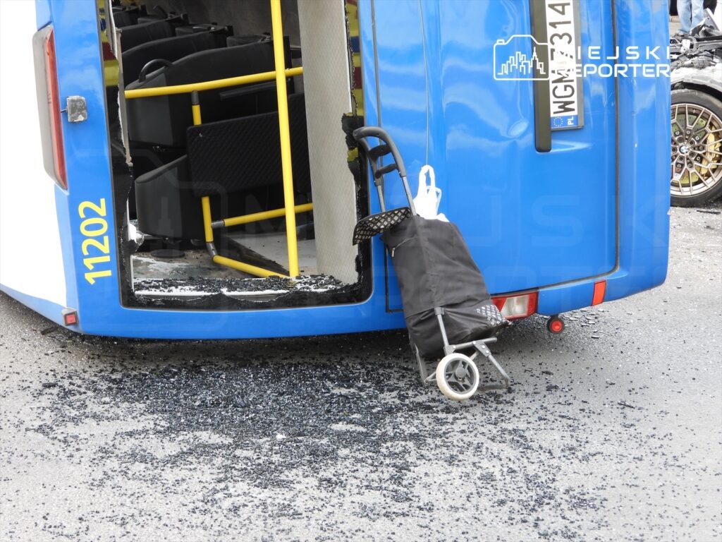 Przewrócony niebieski autobus z odsłoniętym wnętrzem, obok leży czarny wózek dziecięcy, a na drodze widać kawałki asfaltu.