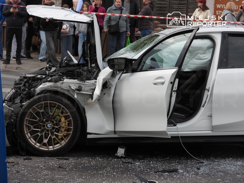 Zniszczony samochód osobowy z otwartymi drzwiami, otoczony przez gapiów, na ulicy po wypadku drogowym.