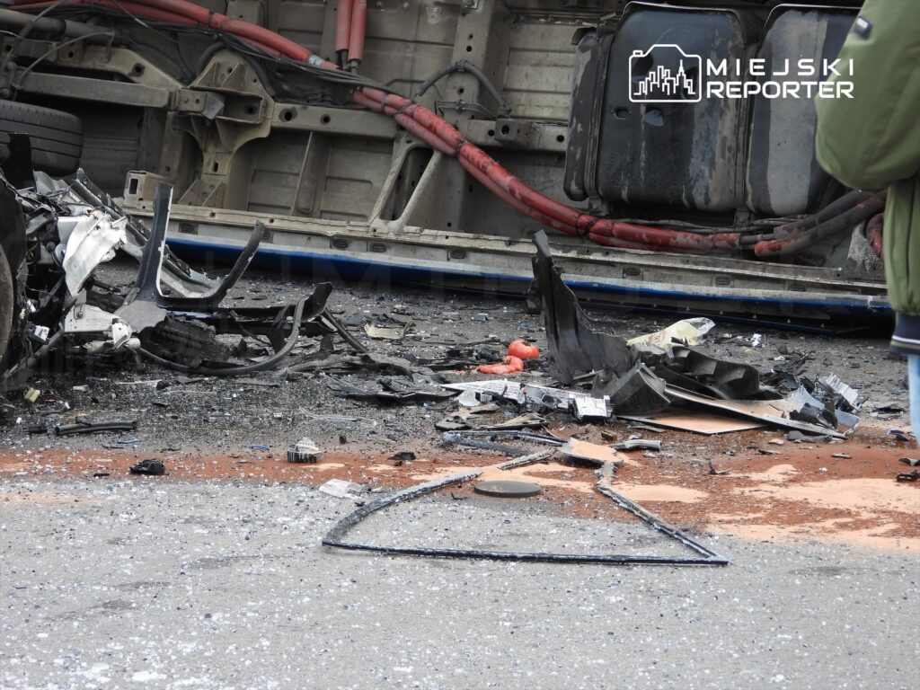Zniszczony wrak pojazdu leżący na jezdni, otoczony fragmentami blachy i innych części, z widocznymi śladami uszkodzeń.