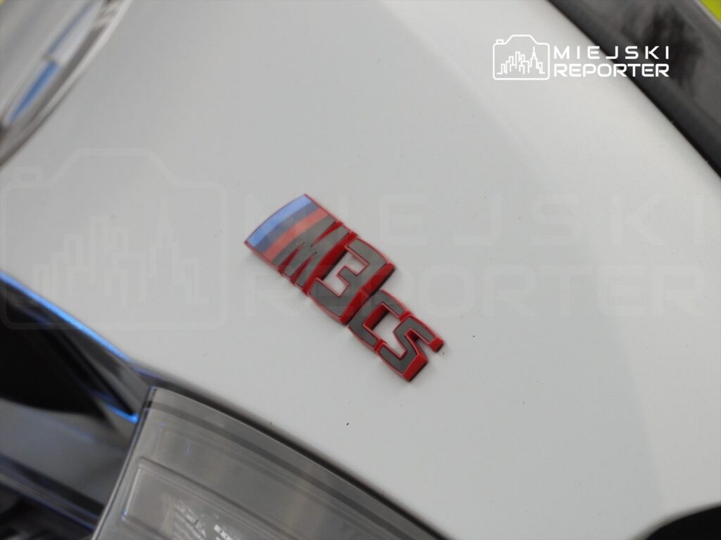 Czerwony emblemat "M3" na białym samochodzie sportowym.
